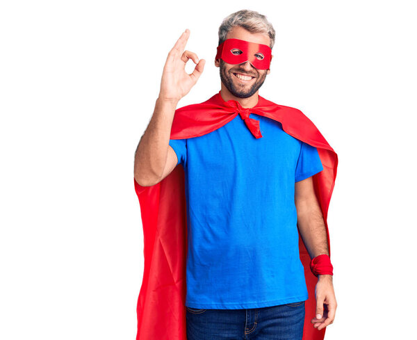 Young blond man wearing super hero custome smiling positive doing ok sign with hand and fingers. successful expression. 
