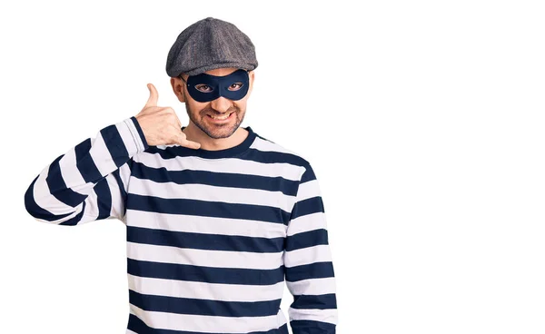 泥棒マスクをした若いハンサムな男が電話で話すような手や指で携帯電話のジェスチャーをして笑っている コミュニケーションの概念 — ストック写真