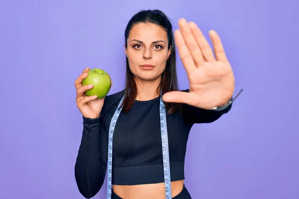テープを使用して美しいブルネットのスポーティな女性は 真剣で自信を持って表現 防衛ジェスチャーで停止記号を行うオープン手で紫色の背景の上にリンゴの果実を食べる測定 — ストック写真