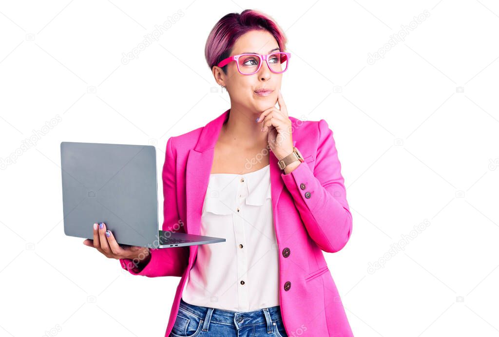 Young beautiful woman with pink hair wearing glasses holding laptop serious face thinking about question with hand on chin, thoughtful about confusing idea 