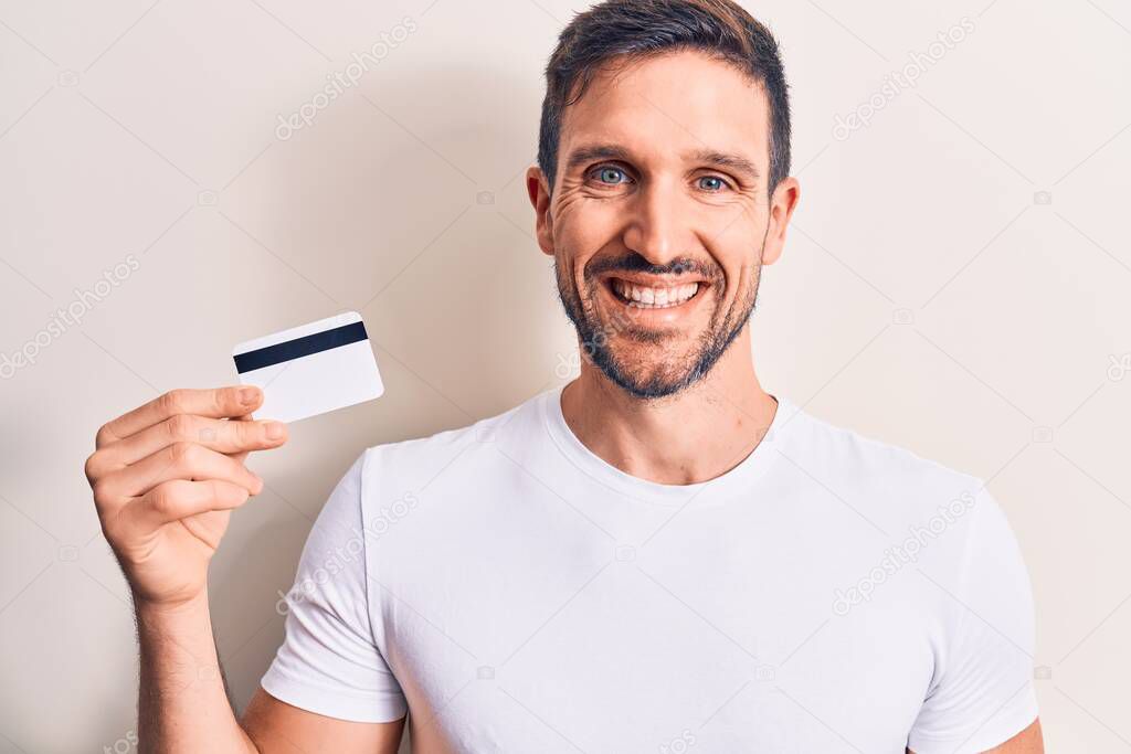 Young handsome customer man holding credit card over isolated white background looking positive and happy standing and smiling with a confident smile showing teeth