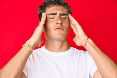 Genç ve yakışıklı bir adam, günlük beyaz tişört giyiyor, elleri başının üstünde, stresten baş ağrısı çekiyor. migren ağrısı. 