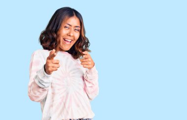 Genç, güzel, karışık ırktan, günlük kravat boyalı kazak giyen, mutlu ve komik yüzlü, parmaklarını kameraya gösteren bir kadın. iyi enerji ve titreşimler. 