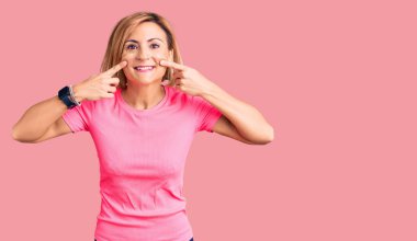 Spor giyinen genç sarışın kadın ağzı açık gülümsüyor, parmakları işaret ediyor ve zorla neşeli bir gülümseme sergiliyor. 