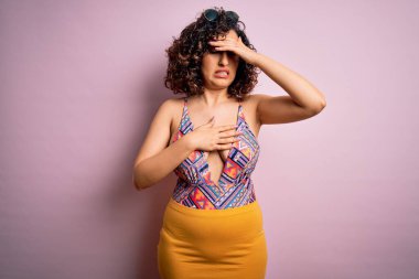 Genç, güzel bir Arap kadın mayo ve güneş gözlüğü takmış pembe arka planda hastalık, ateş, grip ve soğuk algınlığı için alnına dokunuyor.