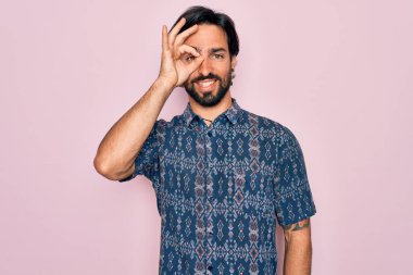 Pembe arka planda hippi tarzı giyinmiş genç, İspanyol bohem bir adam. El hareketleriyle gülümsüyor, gözleriyle gülen parmaklarının arasından bakıyor..