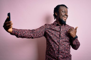 Genç, yakışıklı, Afro-Amerikan bir adam, günlük gömleğini giymiş, kamerayla selfie çekiyor ve gülümseyen bir gülümsemeyle baş parmağıyla onu işaret ediyor.