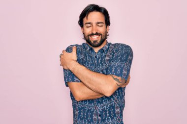 Genç, yakışıklı, bohem, pembe arka planda hippi tarzı giyinmiş mutlu, pozitif ve kendinden emin bir şekilde kendine sarılan bir adam. Sevgi ve özgüven.