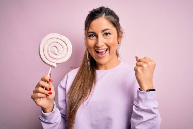 Genç, güzel esmer kadın pembe arka planda şeker yerken gururla bağırıyor, zaferi ve başarıyı kutluyor. Çok heyecanlı, coşkulu bir duygu.