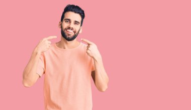 Sakallı, sıradan tişörtlü, neşeli gülümseyen ve parmaklarıyla ve ağzıyla işaret eden genç yakışıklı bir adam. diş sağlığı kavramı. 