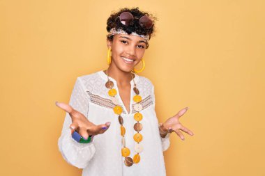 Young beautiful african american afro hippie woman wearing sunglasses and accessories smiling cheerful with open arms as friendly welcome, positive and confident greetings clipart