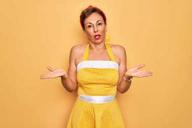 Orta yaşlı, son sınıf rozetli, 50 'li yaşlarda, sarı arka planda retro giysili bir kadın. Hiçbir şeyden habersiz ve kolları açık, hiçbir fikri yok..