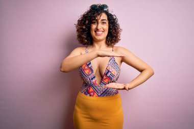 Tatilde olan genç ve güzel bir Arap kadın mayo ve pembe arka planda güneş gözlüğü takıyor. Büyük ve büyük beden işareti, ölçü sembolü gösteren el hareketleriyle. Kameraya bakıp gülümsüyor. Ölçüm konsepti.