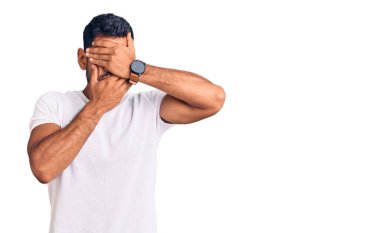 Gündelik kıyafetler giyen genç latin adam gözlerini ve ağzını elleriyle kapatıyor, şaşırmış ve şok olmuş. duygu gizleme 