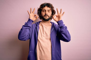 Genç, yakışıklı, sakallı, pembe arka planda sıradan bir kazak giyen, rahatlamış, gözleri kapalı, parmaklarıyla meditasyon hareketi yapan yakışıklı bir adam. Yoga konsepti.