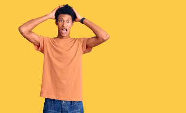 Genç Afrikalı Amerikalı sıradan kıyafetler giyen çılgın ve elleri başında korkmuş ağzı açık bir şekilde şok geçirmekten korkan ve şaşıran bir adam. 