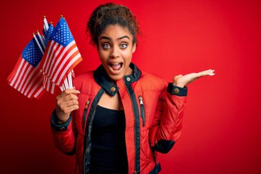 Genç Afro-Amerikan vatansever kız izole edilmiş kırmızı arka planda bayraklar tutuyor. Mutlu ve heyecanlı, kazanan ifade zaferi büyük bir gülümsemeyle kutluyor ve ellerini kaldırıyor.