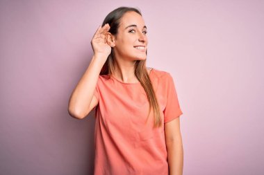 İzole edilmiş pembe arka planda dikilmiş, rahat bir tişört giyen genç ve güzel bir kadın kulağına fısıldayarak dedikoduları ve dedikoduları dinliyor. Sağırlık kavramı.