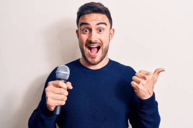 Sakallı genç yakışıklı şarkıcı beyaz arka planda mikrofon kullanarak şarkı söylüyor baş parmağını yukarı kaldırıyor ve ağzı açık gülümsüyor.