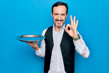 Orta yaşlı, yakışıklı garson adam mavi arka planda duruyor. Tamam işareti yapıyor. Parmaklarıyla, gülümseyerek mükemmel bir sembol oluşturuyor.