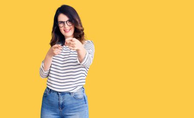 Güzel, genç, esmer, sıradan kıyafetler giyen ve gözlüklü bir kadın mutlu ve komik yüzüyle kamerayı işaret ediyor. iyi enerji ve titreşimler. 