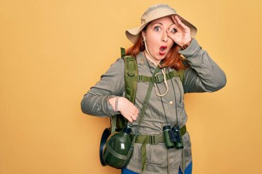 Sırt çantası ve sarı arka planda şapka takan kızıl saçlı genç bir kadın şaşırmış yüz ifadesiyle şaşırmış ve parmaklarından bakan bir jest yapıyor. İnançsız ifade.