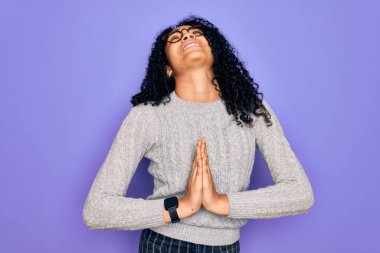 Genç Afro-Amerikalı kadın mor arka planda günlük süveter ve gözlük takıyor. Elleriyle yalvarıyor ve dua ediyor. Yüzünde umut ifadesi var. Çok duygusal ve endişeli. Yalvarıyorum..