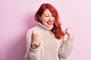 Genç, güzel, kızıl saçlı kadın pembe arka plan üzerine balıkçı yaka kazak giyiyor. Şaşırmış ve şaşırmış bir şekilde kolları havada ve gözleri kapalı bir şekilde başarıyı kutluyor.