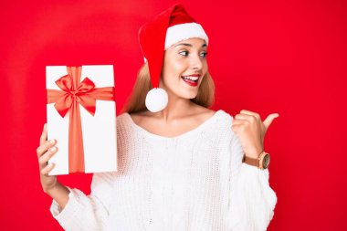 Genç esmer kadın, Noel şapkası takıyor ve elinde bir hediye var. Parmağını kaldırıp gülümsüyor ve ağzı açık. 