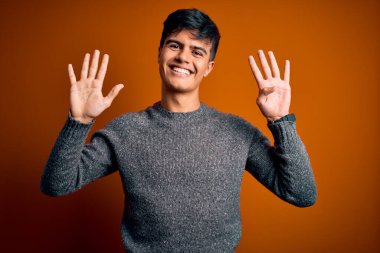 İzole edilmiş turuncu arka plan üzerinde duran sıradan süveter giyen genç ve yakışıklı bir adam dokuz numaralı parmaklarıyla işaret ederken kendinden emin ve mutlu bir şekilde gülümsüyor..