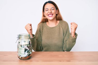 Genç esmer kadın, para dolu bir kavanozla masada oturuyor çok mutlu ve heyecanlı bir şekilde kollarını kaldırıp, gülümseyerek ve başarı için çığlık atarak zafer jesti yapıyor. kutlama konsepti. 