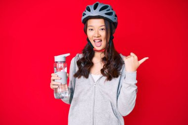 Bisiklet kaskı takan ve elinde su şişesi tutan genç, güzel Çinli bir kız başparmağıyla yan tarafa doğru gülümsüyor ve ağzı açık. 