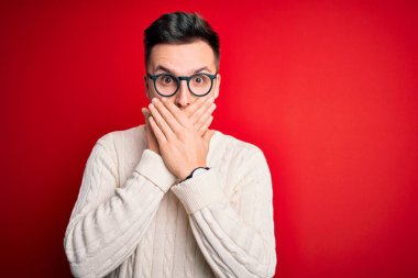 Genç, beyaz, gözlüklü ve kırmızı arka planda günlük kışlık süveter giyen yakışıklı bir adam ağzını yanlışlıkla ellerle kapatarak şok etti. Gizli konsept.