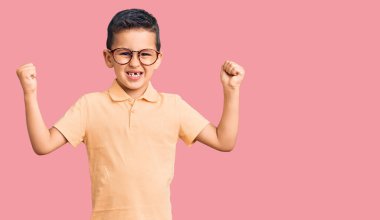 Küçük sevimli çocuk günlük kıyafetler ve gözlük takıyor gururla bağırıyor, zaferi kutluyor ve kollarını kaldırarak başarıyı kutluyor. 