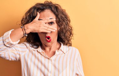 Orta yaşlı, esmer, çizgili tişört giyen güzel bir kadın sarı arka planda dikiliyor. Şok olmuş yüzünü ve gözlerini eliyle kapatıyor, parmaklarından korkuyla bakıyor.