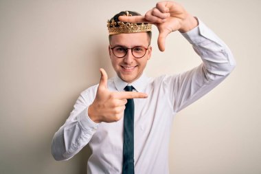 İzole edilmiş arka planda altın taç takan yakışıklı beyaz iş adamı gülümseyerek elleri ve parmakları gülen yüzlü bir çerçeve yapıyor. Yaratıcılık ve fotoğrafçılık kavramı.