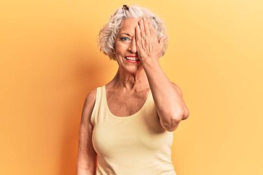 Kıdemli gri saçlı kadın, günlük kıyafetler giyiyor, bir gözünü eliyle kapatıyor, yüzünde kendine güvenen bir gülümseme ve şaşırtıcı bir duygu var.. 