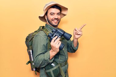 Kaşif kıyafetleri giyen genç İspanyol adam elinde dürbünle mutlu bir şekilde gülümsüyor. 