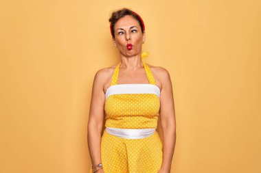 Orta yaşlı, 50 'li yaşlarda, sarı arka plan üzerine eski moda elbise giyen, dudağıyla balık suratı yapan, çılgın ve komik bir jest yapan bir kadın. Komik ifade.