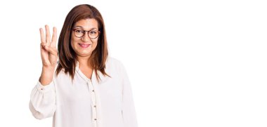 Orta yaşlı Latin kadın gündelik kıyafetler ve gözlük takıyor. Üç numaralı parmaklarıyla işaret ediyor ve gülümsüyor. Kendinden emin ve mutlu.. 