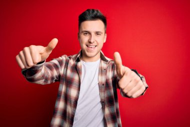 Kırmızı izole edilmiş arka plan üzerine gündelik modern tişört giyen genç beyaz adam elle olumlu jest yapmayı onaylıyor, gülümsüyor ve başarı için mutlu. Kazanan jest.