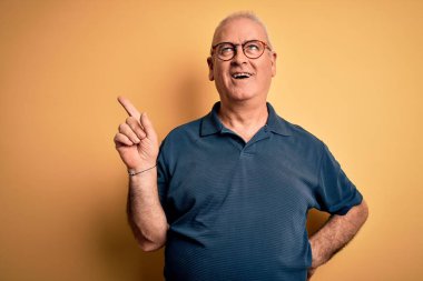 Orta yaşlı, beyaz tenli, sıradan polo giyen ve sarı arka planda gözlüklü, yüzünde kocaman bir gülümseme olan yakışıklı bir adam, kameraya el parmağı ile bakıyor..