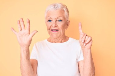 Son sınıf güzeli, mavi gözlü, gri saçlı, sarı arka planda klasik beyaz tişört giyen ve 6 numaralı parmaklarıyla işaret eden, kendinden emin ve mutlu gülümseyen bir kadın.. 