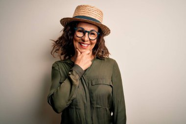 Orta yaşlı esmer kadın, gözlüklü ve şapkalı, izole edilmiş beyaz arka planda duruyor. Kameraya kendinden emin bir şekilde bakıyor. Kollarını çapraz yapıp, elini çenesine kaldırmış gülümsüyor. Olumlu düşünüyorum..