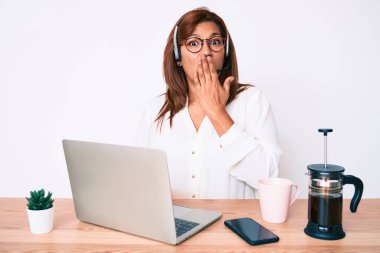 Orta yaşlı esmer kadın ofiste çalışıyor operatör kulaklığı takıyor ağzı elle kapatıyor, şok geçiriyor ve hata yapmaktan korkuyor. şaşırmış ifade 