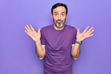 Orta yaşlı, mor tişört giyen yakışıklı bir adam izole edilmiş arka planda duruyor. Hiçbir şey bilmiyor ve kolları açık, hiçbir fikri ve şüpheli bir yüzü var..