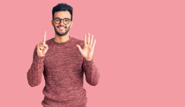 Kış süveteri giyen ve gözlük takan genç yakışıklı İspanyol adam altı numaralı parmaklarını gösterip işaret ederken kendinden emin ve mutlu gülümsüyor.. 