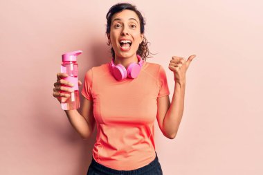 Spor kıyafetleri giyen, kulaklık takan, su içen, ağzı açık gülümseyen genç ve güzel bir İspanyol kadın. 