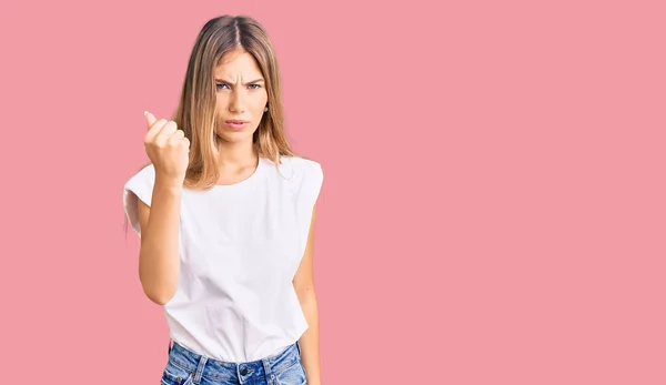 Beautiful caucasian woman with blonde hair wearing casual white tshirt angry and mad raising fist frustrated and furious while shouting with anger. rage and aggressive concept.
