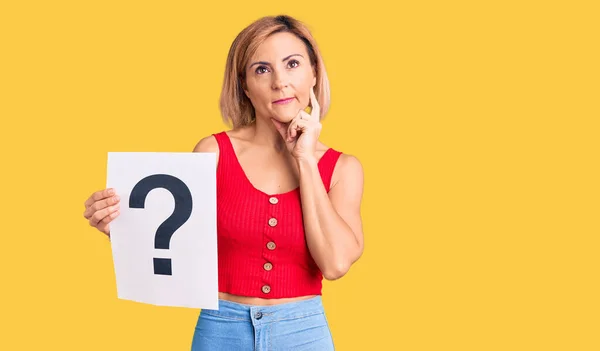 Young blonde woman holding question mark serious face thinking about question with hand on chin, thoughtful about confusing idea